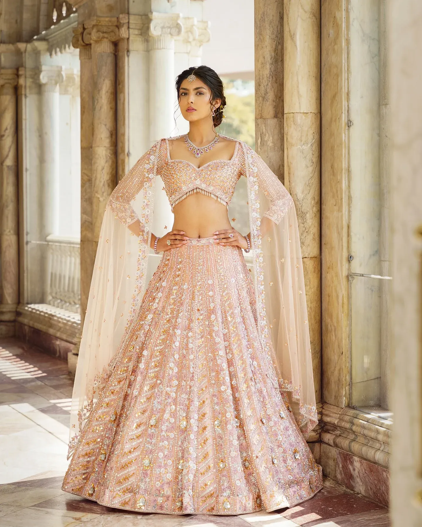 A blush lehenga with sequin embroidery and a sheer cape, beside pale columns.