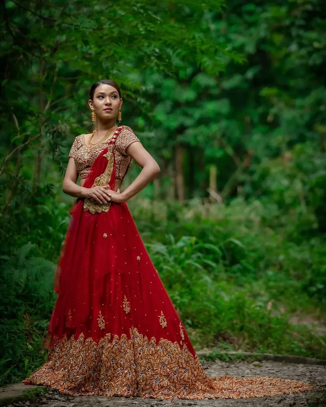 A red lehenga with a wide gold border and a matching dupatta, photographed against deep greenery.