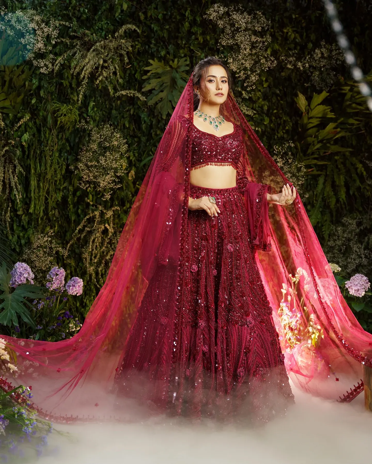 A deep red lehenga with a long veil, photographed among foliage in mist.