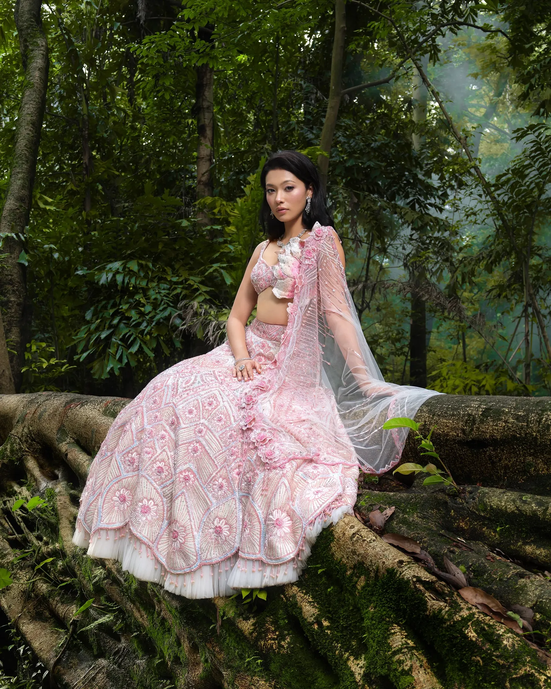 A pink lehenga with ruffled tiers, photographed seated on mossy tree roots.