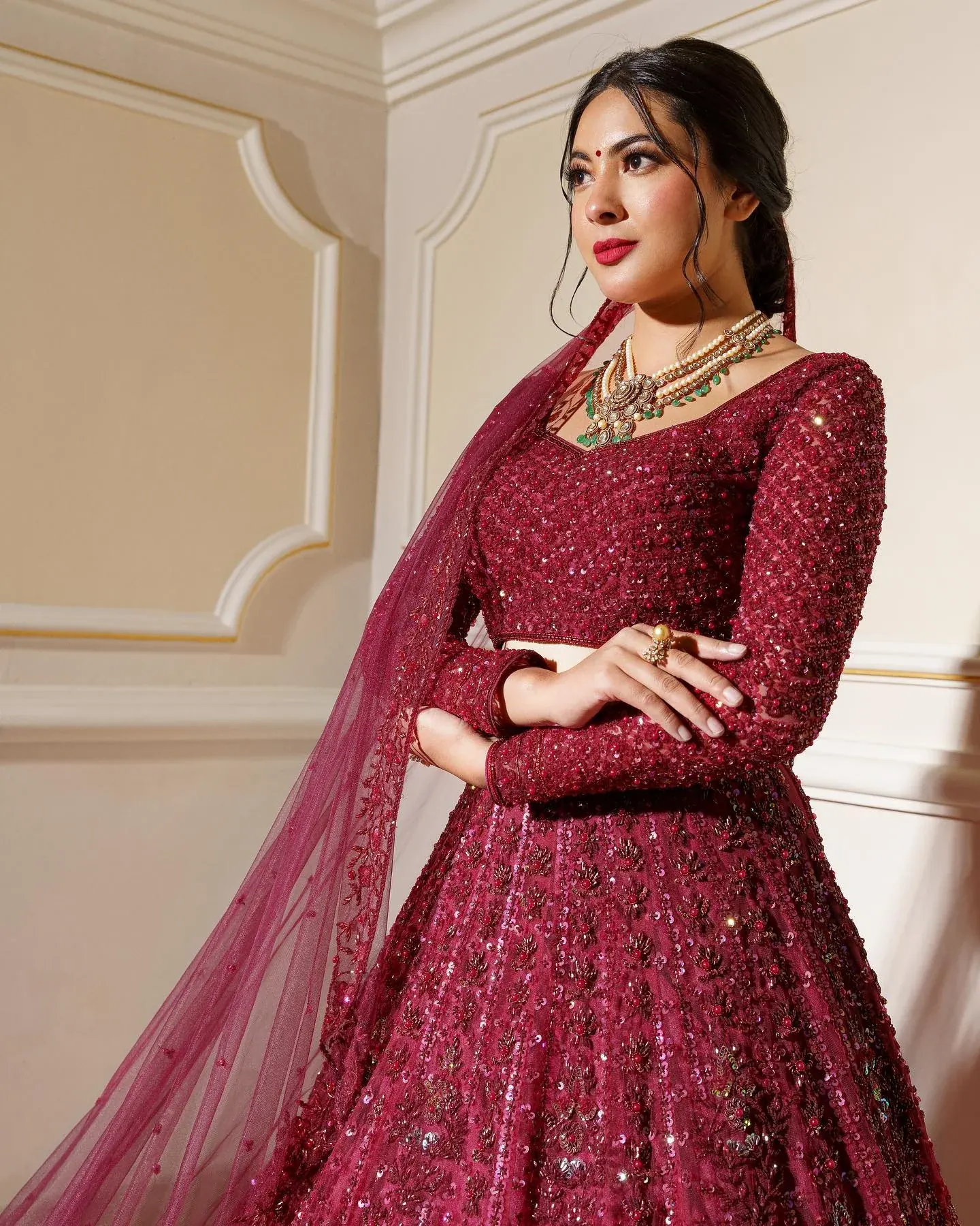 A deep red lehenga with long embroidered sleeves and a sheer dupatta, against a panelled wall.