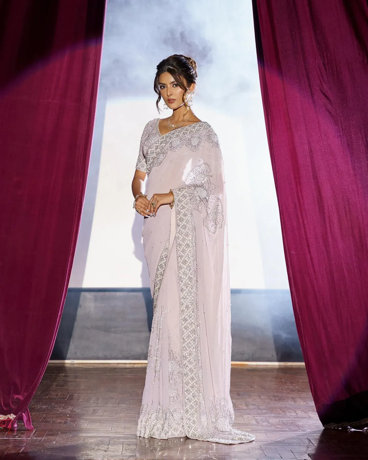A white saree with a scalloped border, photographed on a stage between drawn curtains.