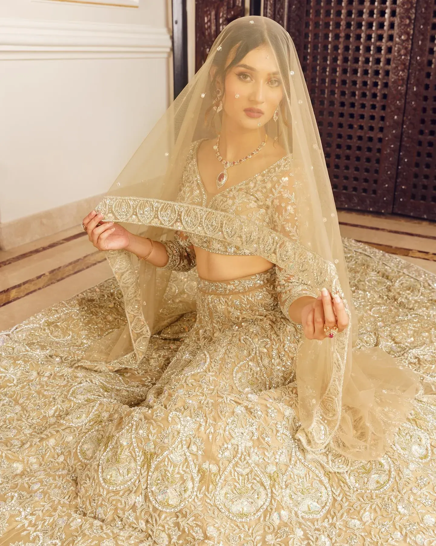 A gold lehenga with dense embroidery and a full veil, seated against a carved screen.