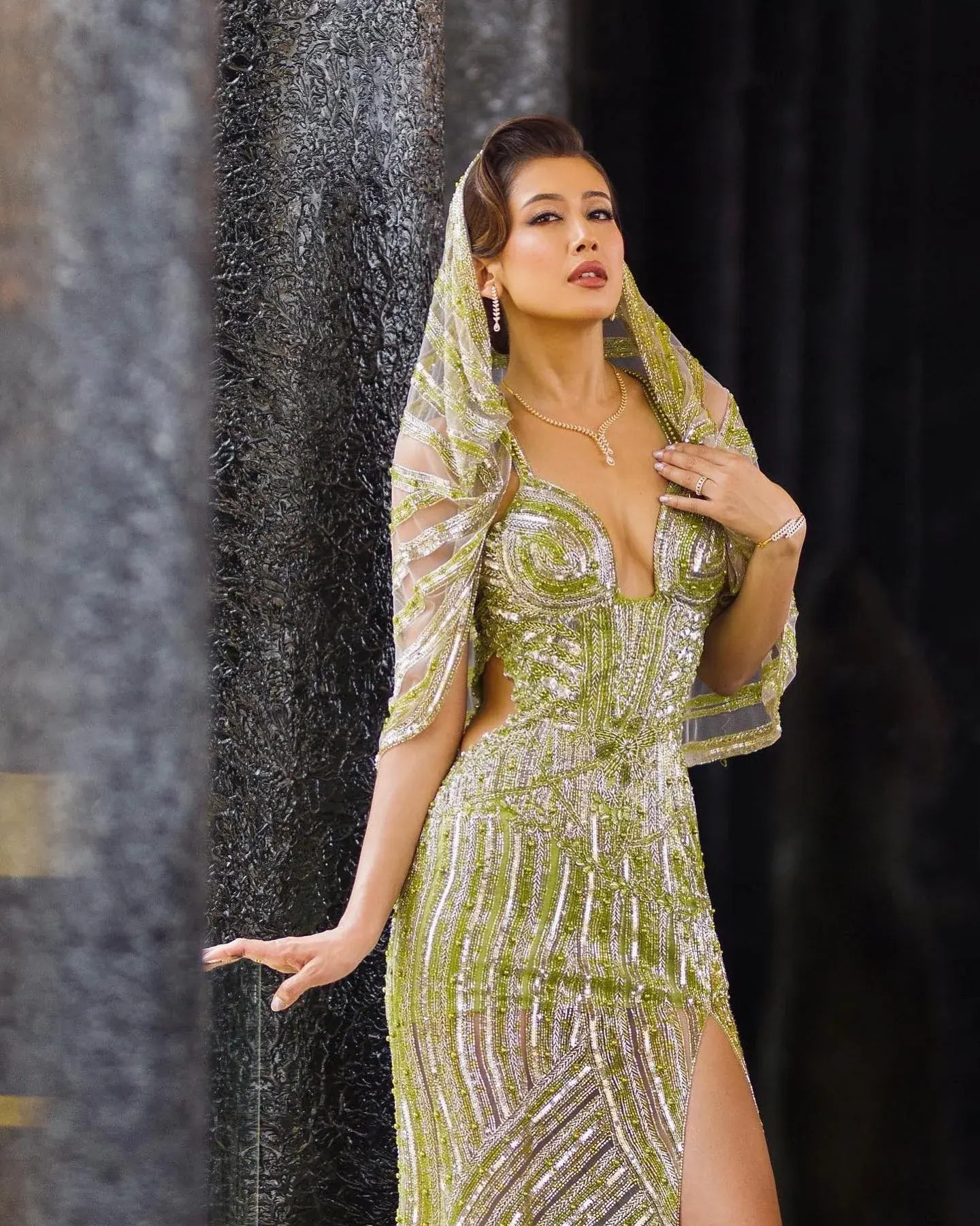 A green and gold beaded gown with a high slit, photographed beside a stone column.