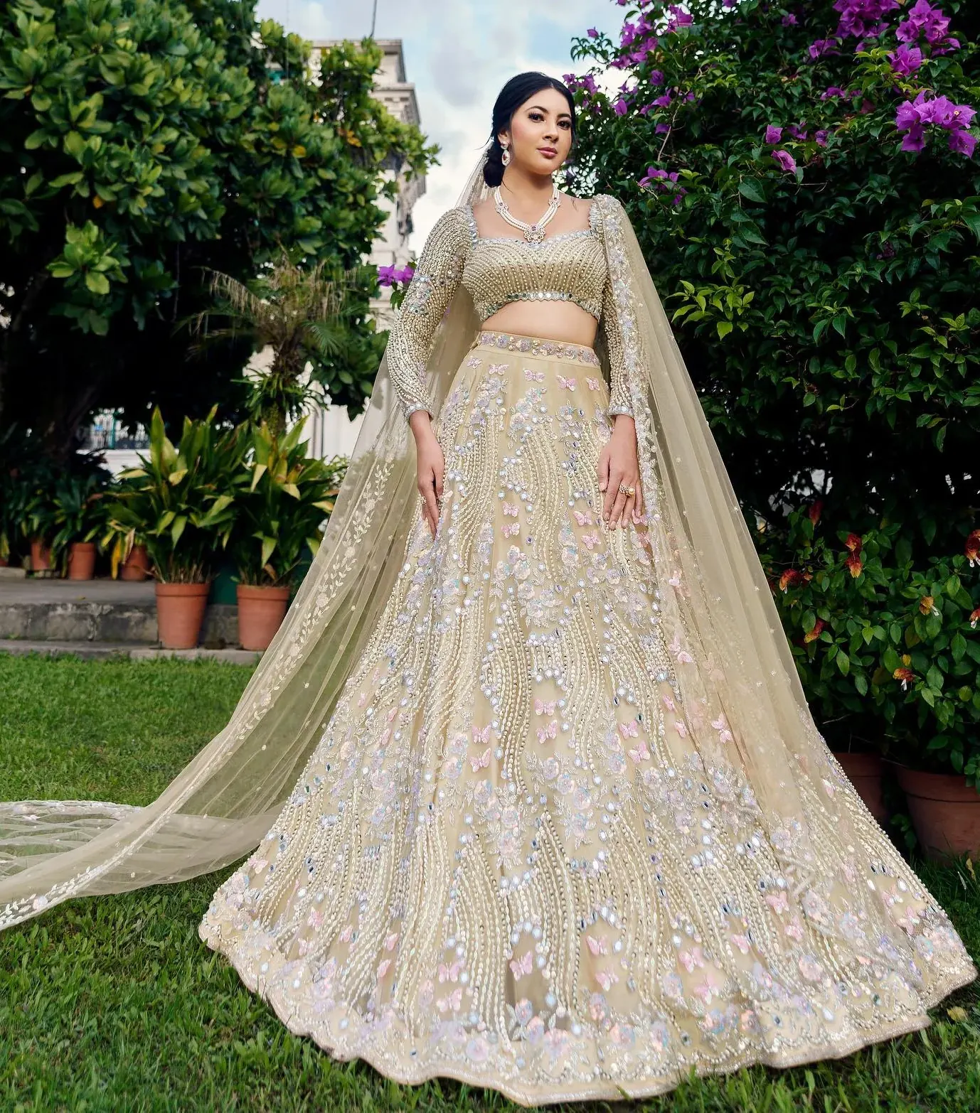 An ivory lehenga with silver thread work and a long sheer dupatta, photographed beside flowering bougainvillea.