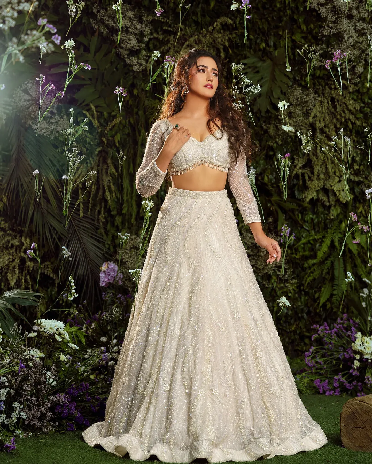 An ivory lehenga with a beaded blouse and sheer sleeves, photographed among foliage.