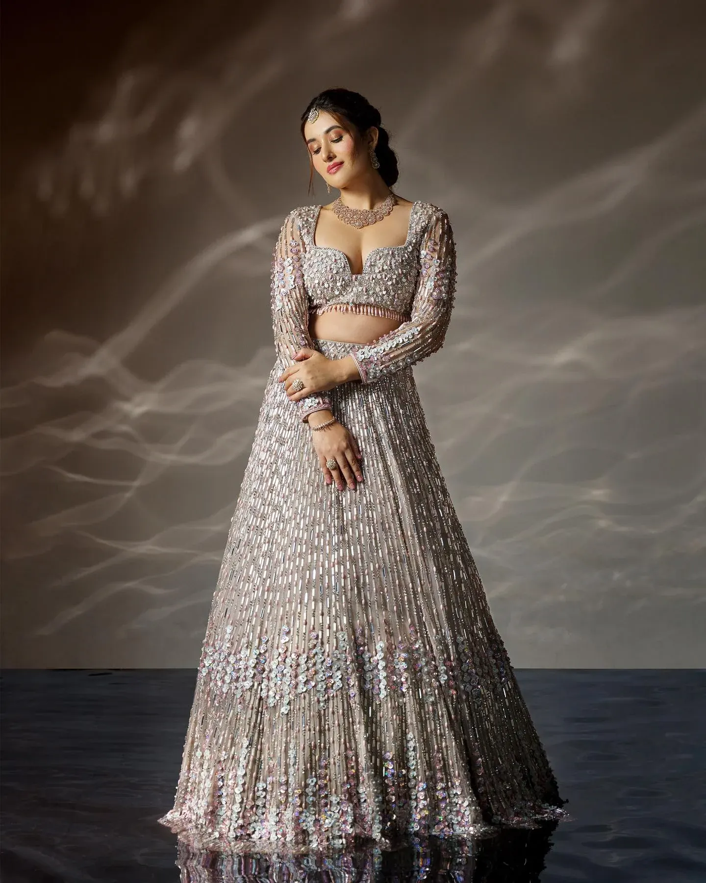 A silver lehenga with dense bead work and long sleeves, against a grey ground.