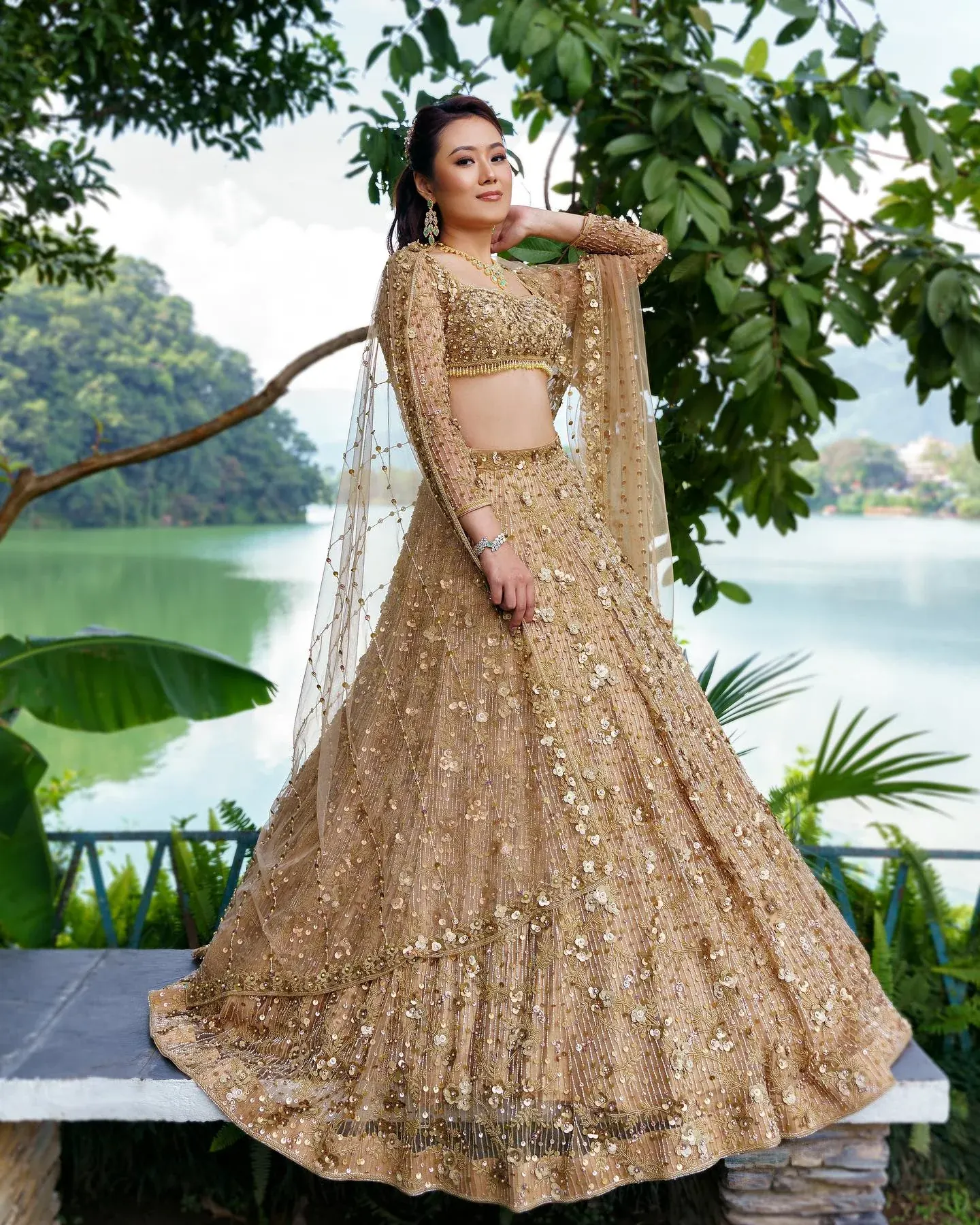 A gold lehenga with dense embroidery and a sheer dupatta, photographed under a tree.