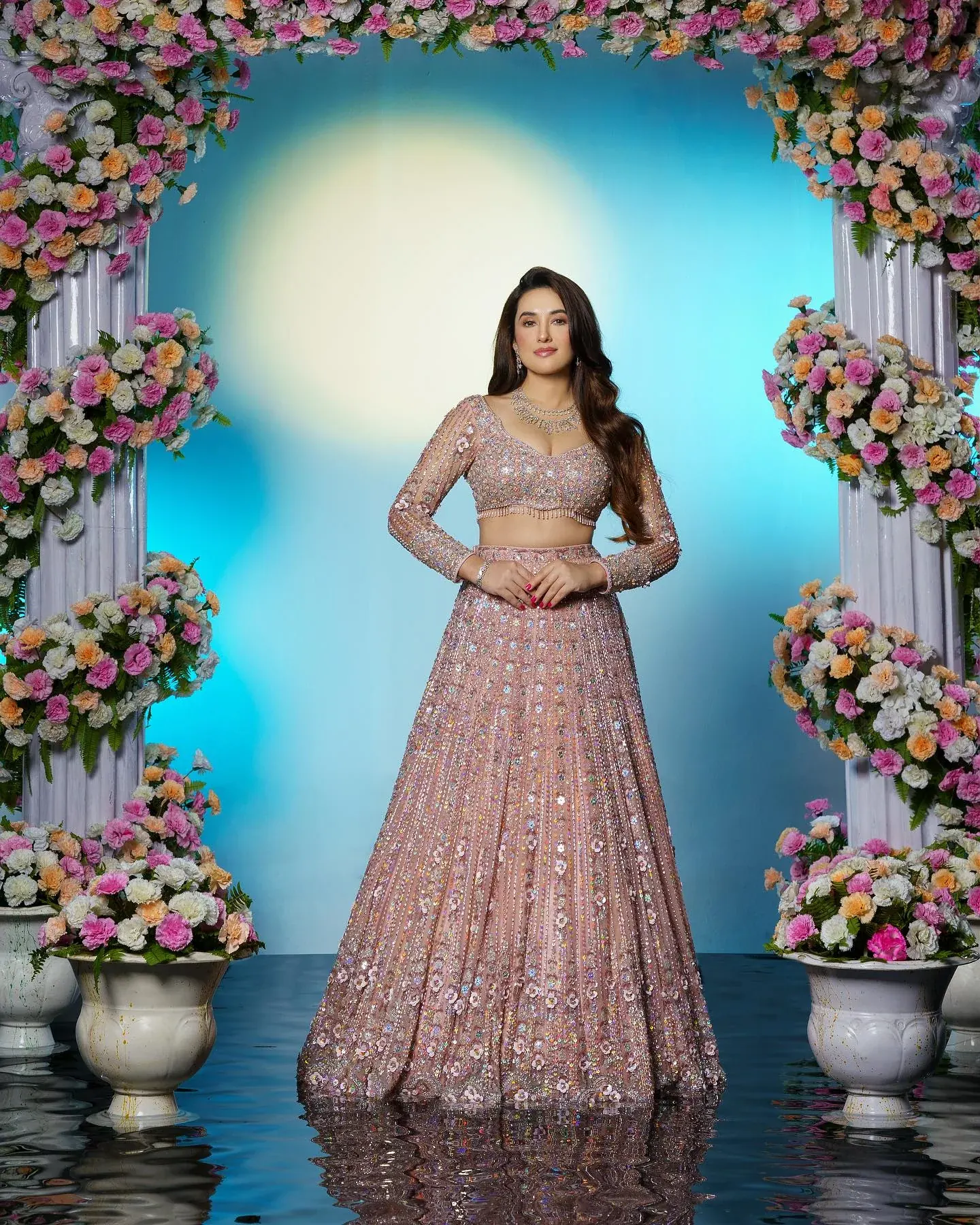 A blush lehenga with silver embroidery, photographed beneath an arch of flowers.
