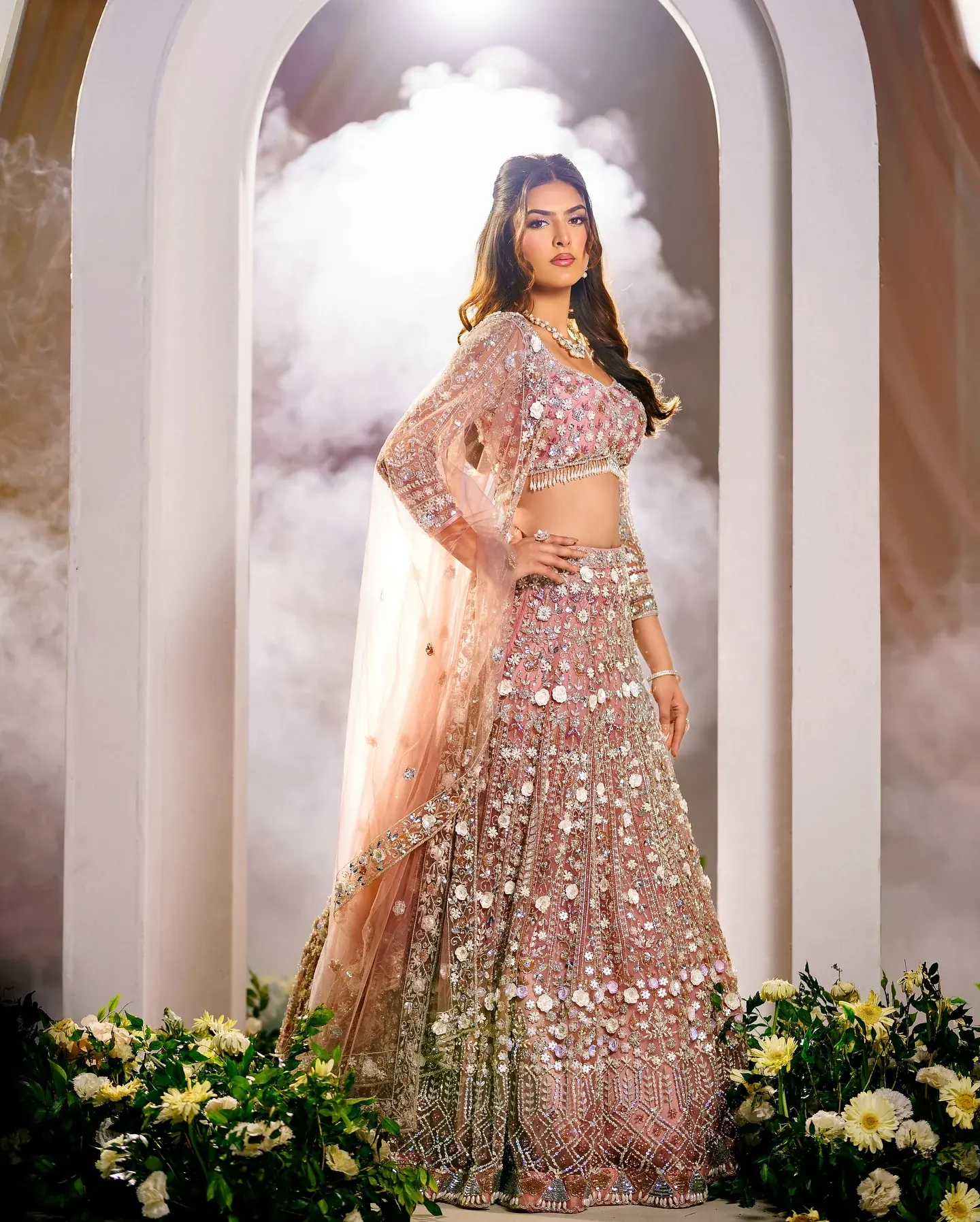 A blush lehenga with silver embroidery, photographed beneath an arch in mist.
