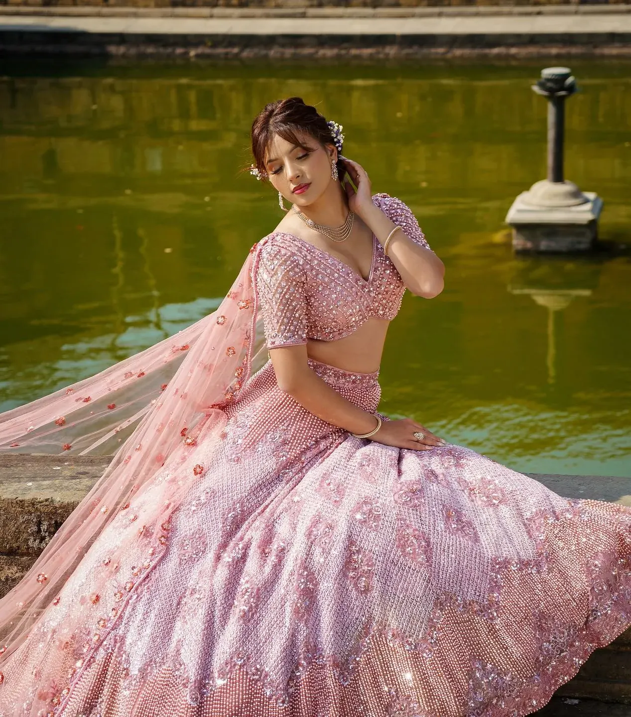 A blush lehenga with a beaded blouse, photographed beside still water.