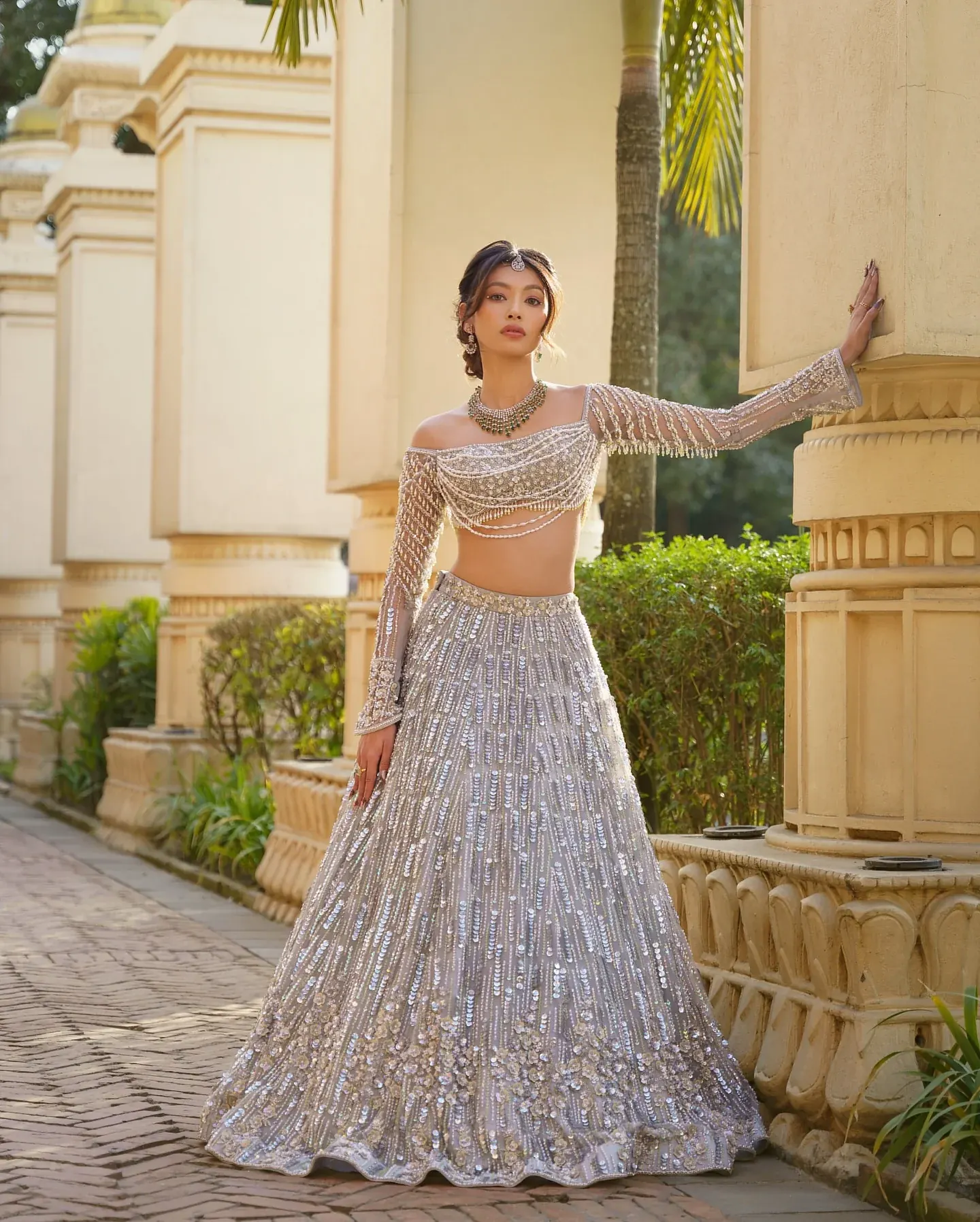 A silver lehenga with dense mirror work and a sheer dupatta, along a colonnade.