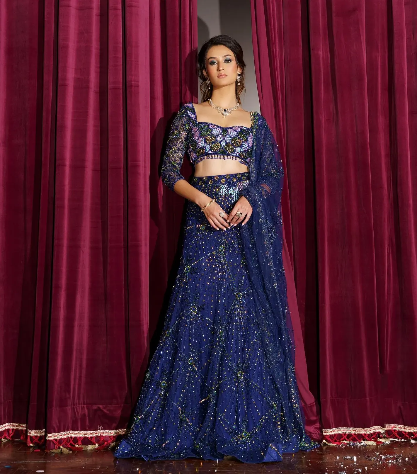 A navy lehenga with sequinned floral work and a beaded blouse, against a red curtain.