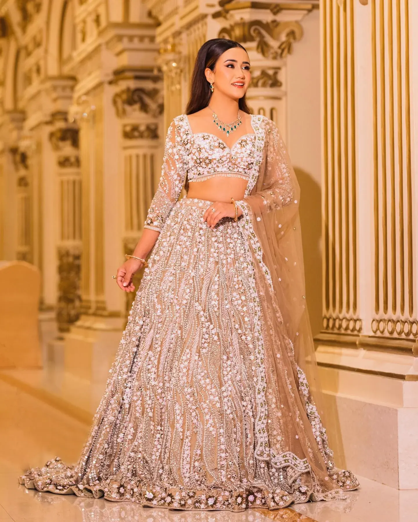 A champagne lehenga with a beaded blouse and sheer dupatta, beside gilded columns.