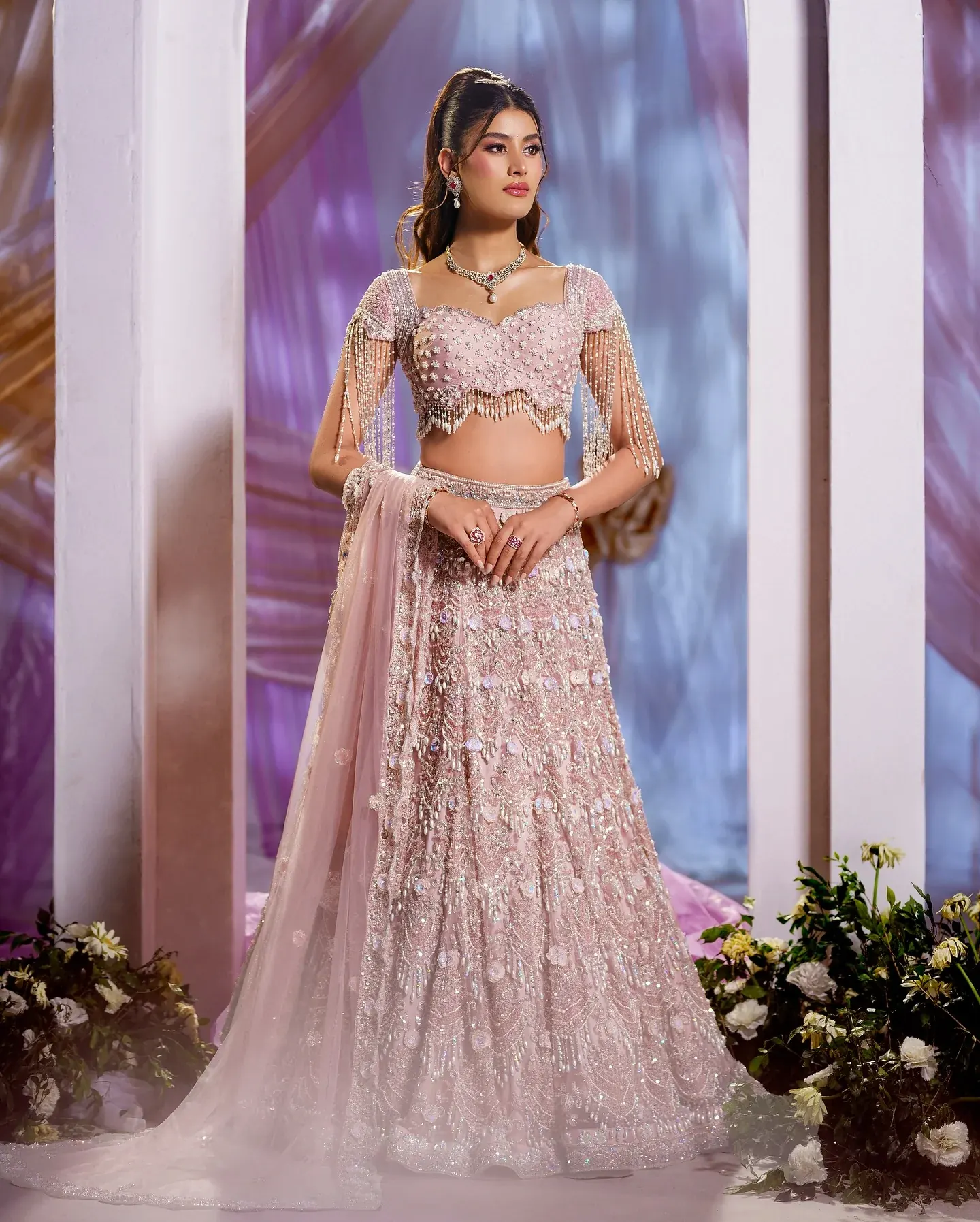 A blush lehenga with a sheer dupatta and beaded blouse, beneath a pale arch.
