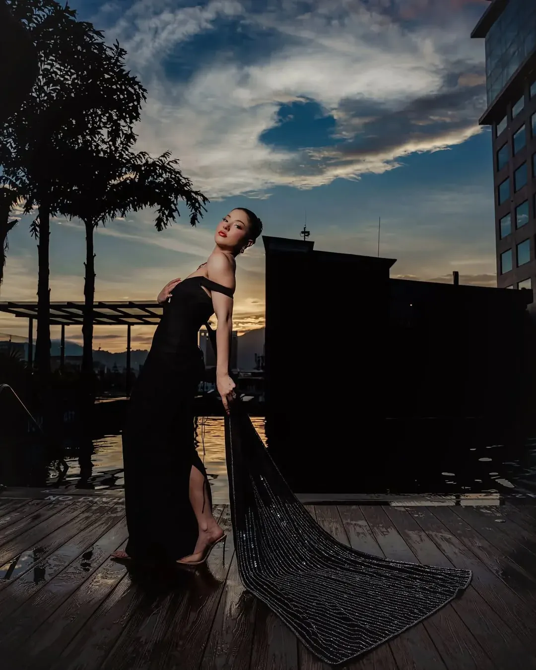 A black gown with a beaded train spread across a wet deck at dusk.