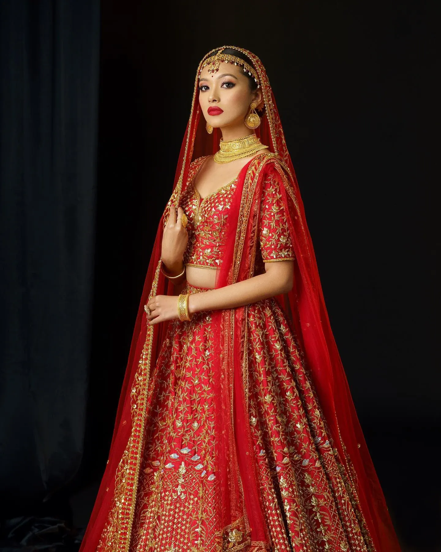 A red bridal lehenga with gold embroidery and a veil, photographed against a dark ground.