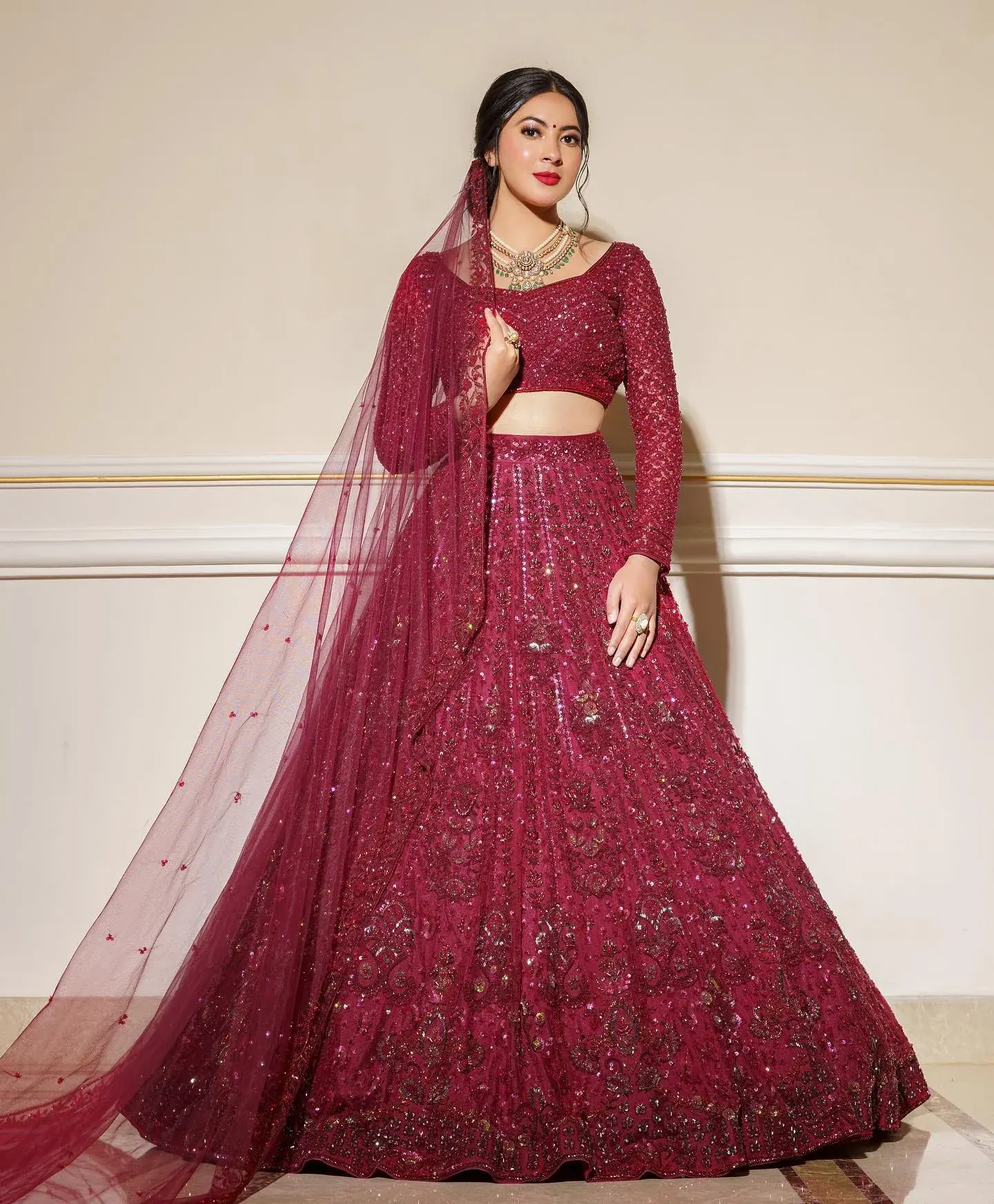 A maroon lehenga with beadwork and a sheer dupatta held wide, against a pale wall.