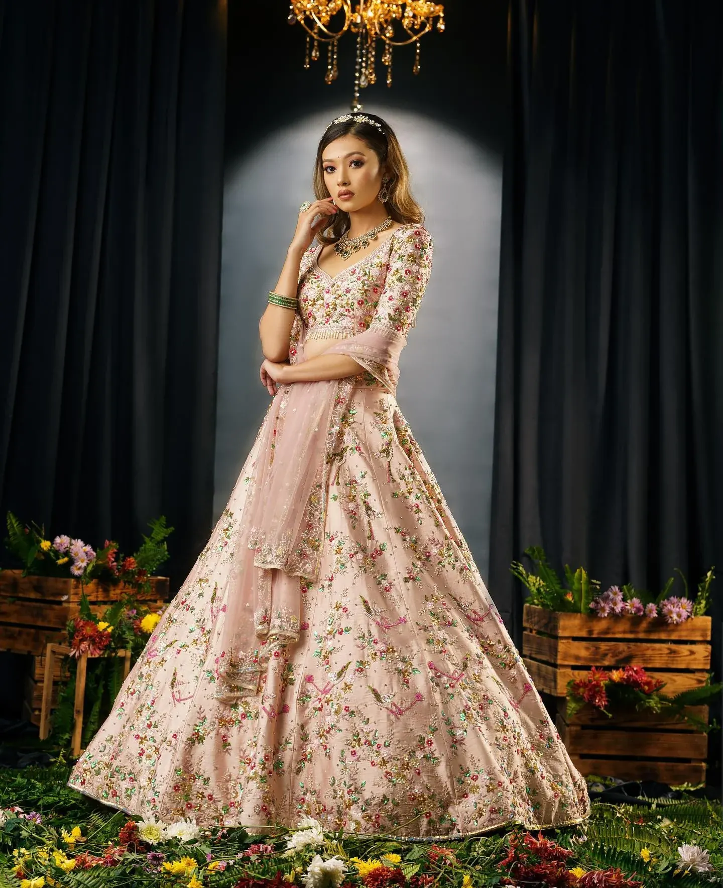 A blush lehenga with floral embroidery, photographed beneath a chandelier among flower crates.
