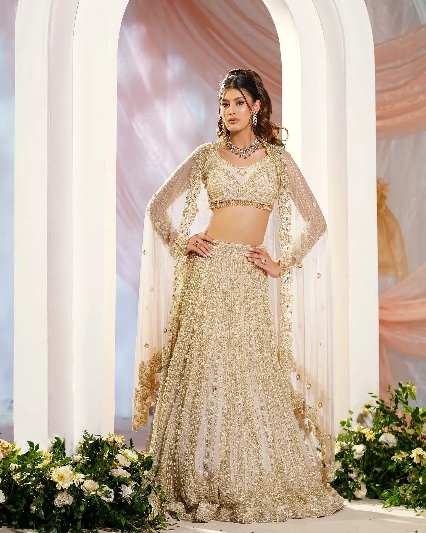 An ivory lehenga with sequin work and a long sheer dupatta, beneath a pale arch.