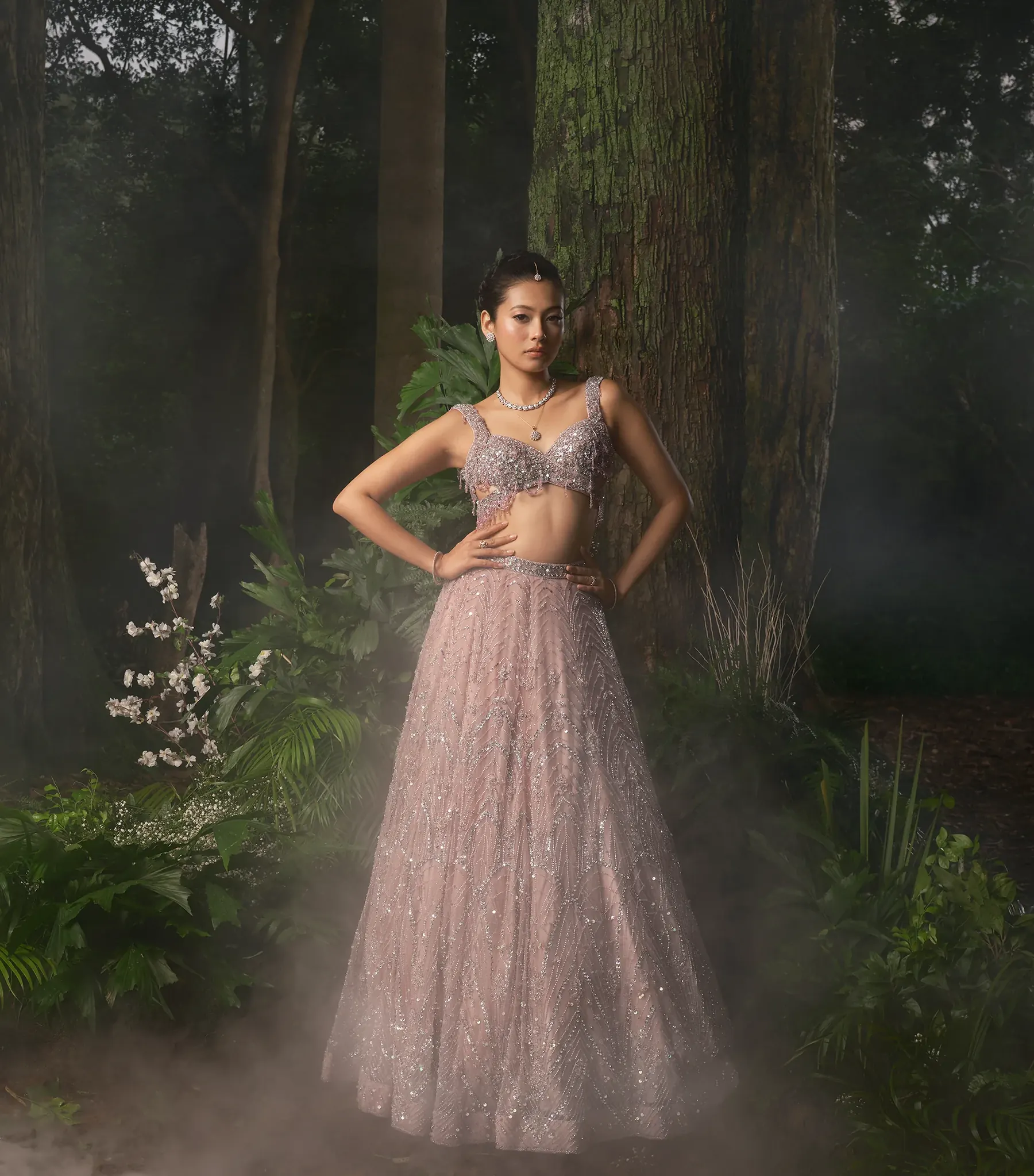 A pale pink lehenga with a beaded bustier blouse and fringe, photographed in mist among dark trees.