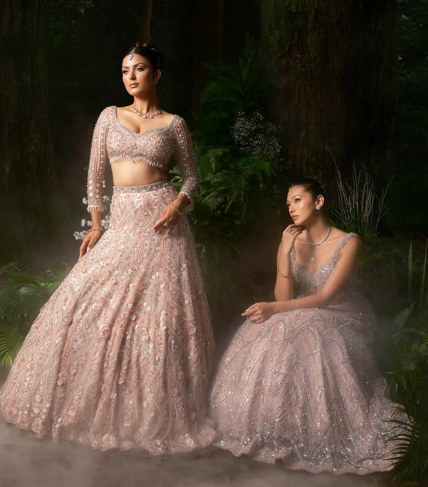 Two pale pink embroidered lehengas photographed together in a misty forest, one standing and one seated.