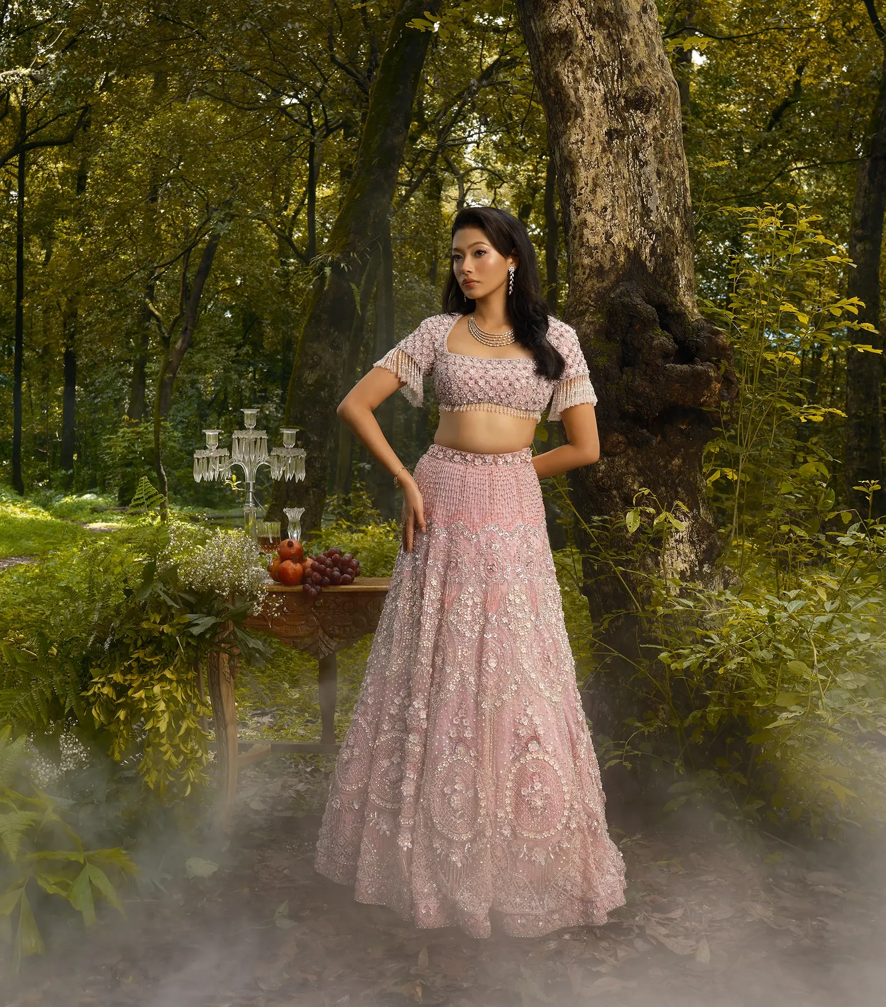 A pale pink lehenga with a scalloped, beaded blouse and tasselled sleeves, beside a carved table and a crystal candelabra.