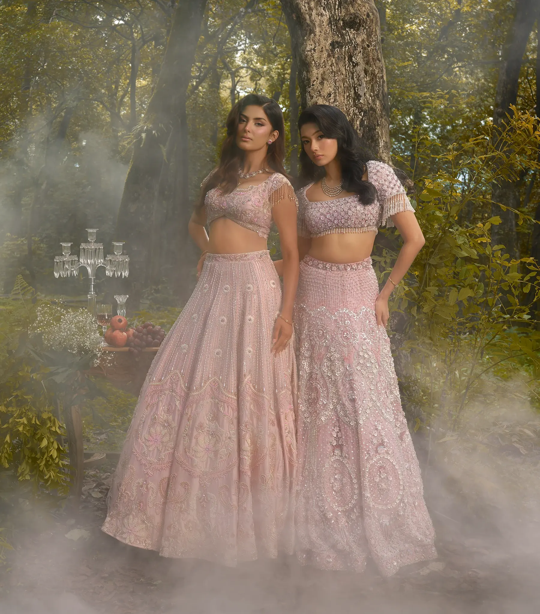 Two blush pink lehengas, one with tasselled sleeves and one with a fringed blouse, photographed together in mist.