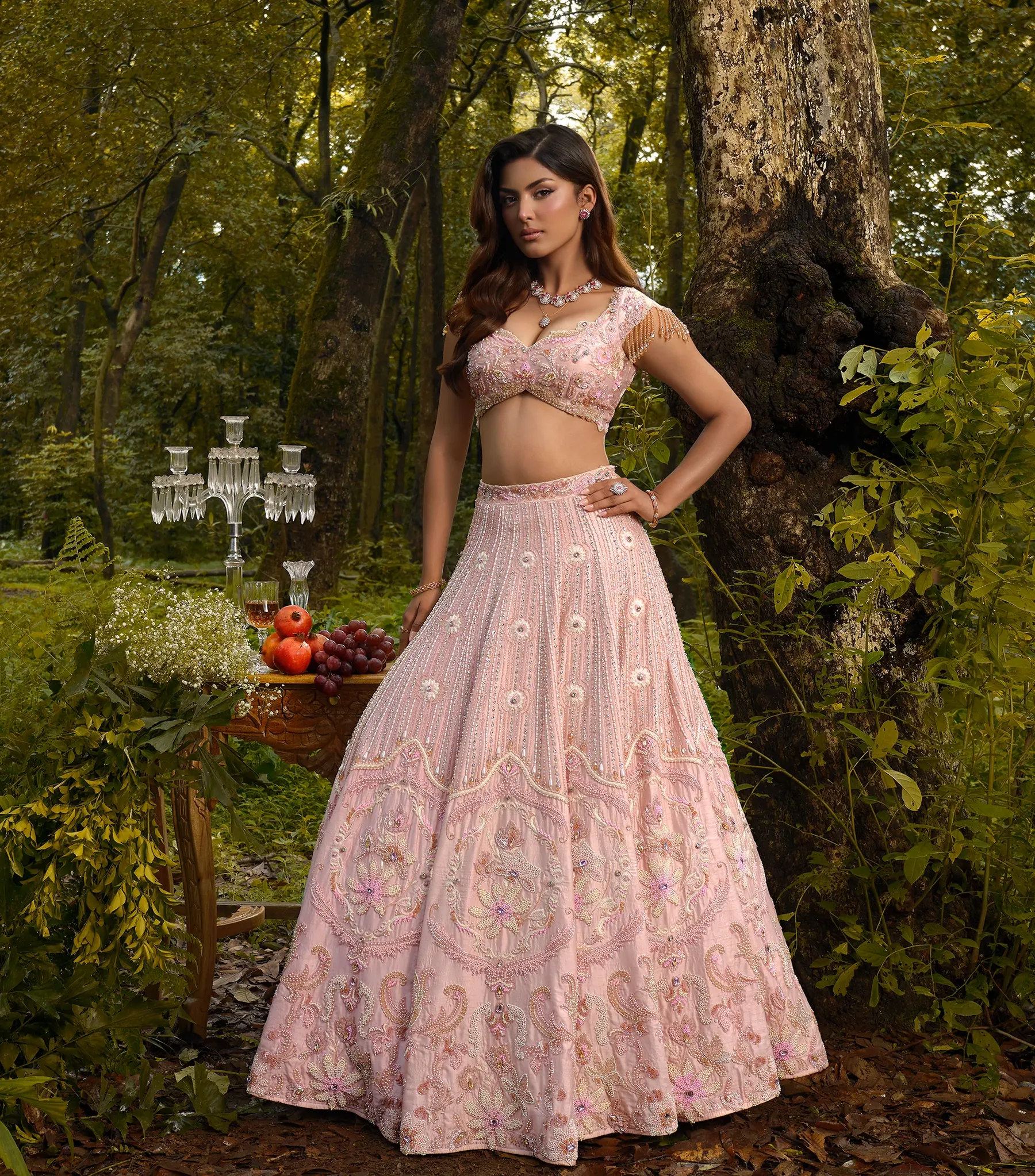A blush pink lehenga with paisley embroidery and a beaded blouse, beside a carved table of fruit and a crystal candelabra.