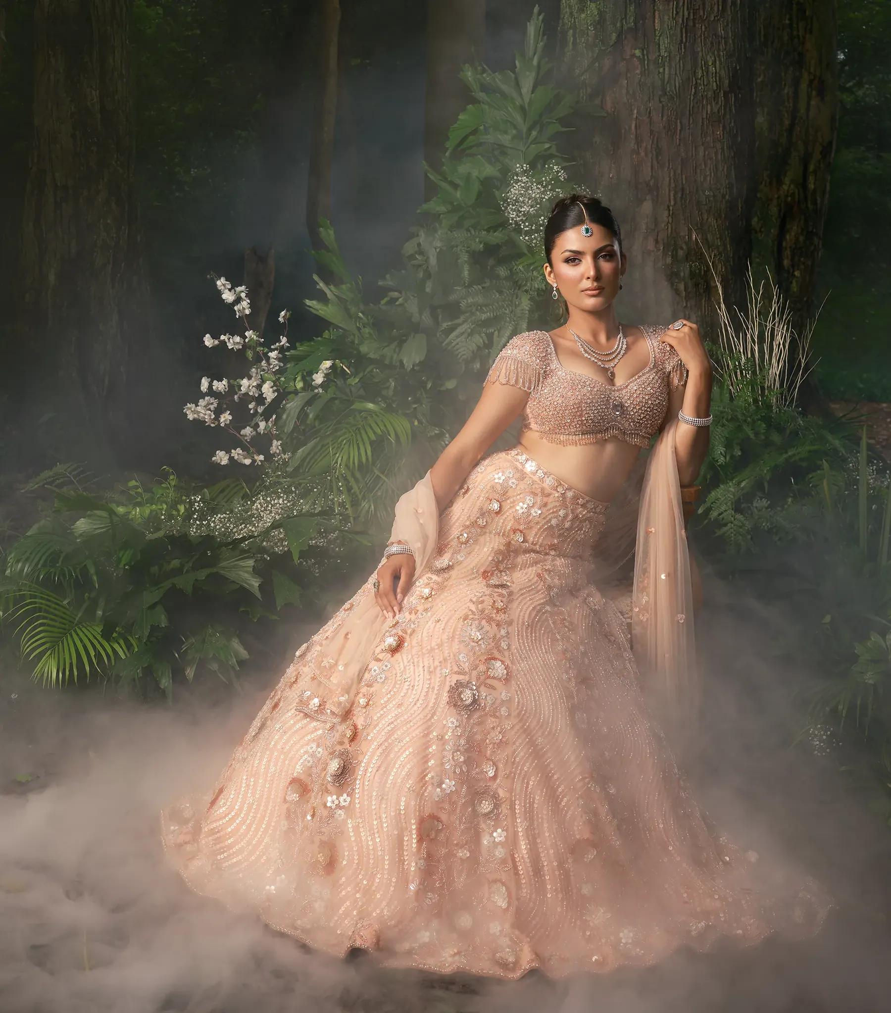 A blush lehenga with floral appliqué and a beaded bralette blouse, photographed beneath a floral arch in mist.