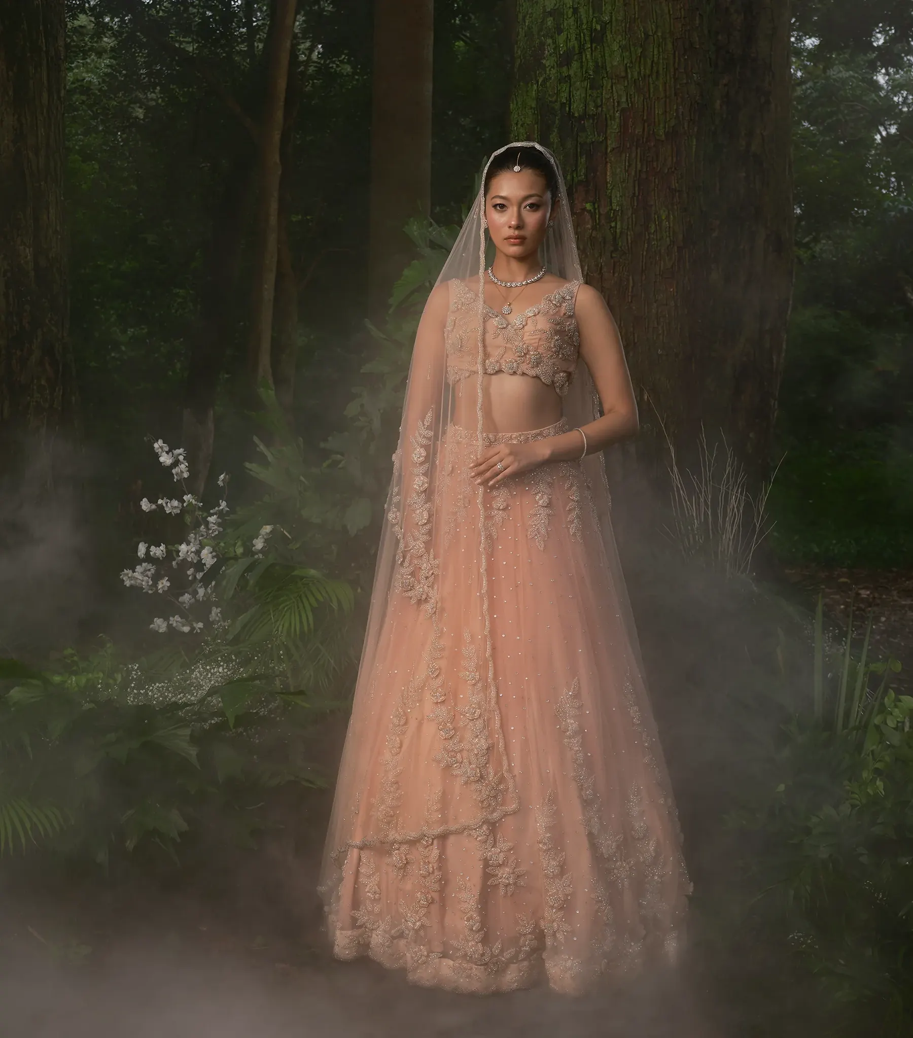 A peach bridal lehenga with a full-length veil and floral appliqué down the drape, in low mist among dark trees.