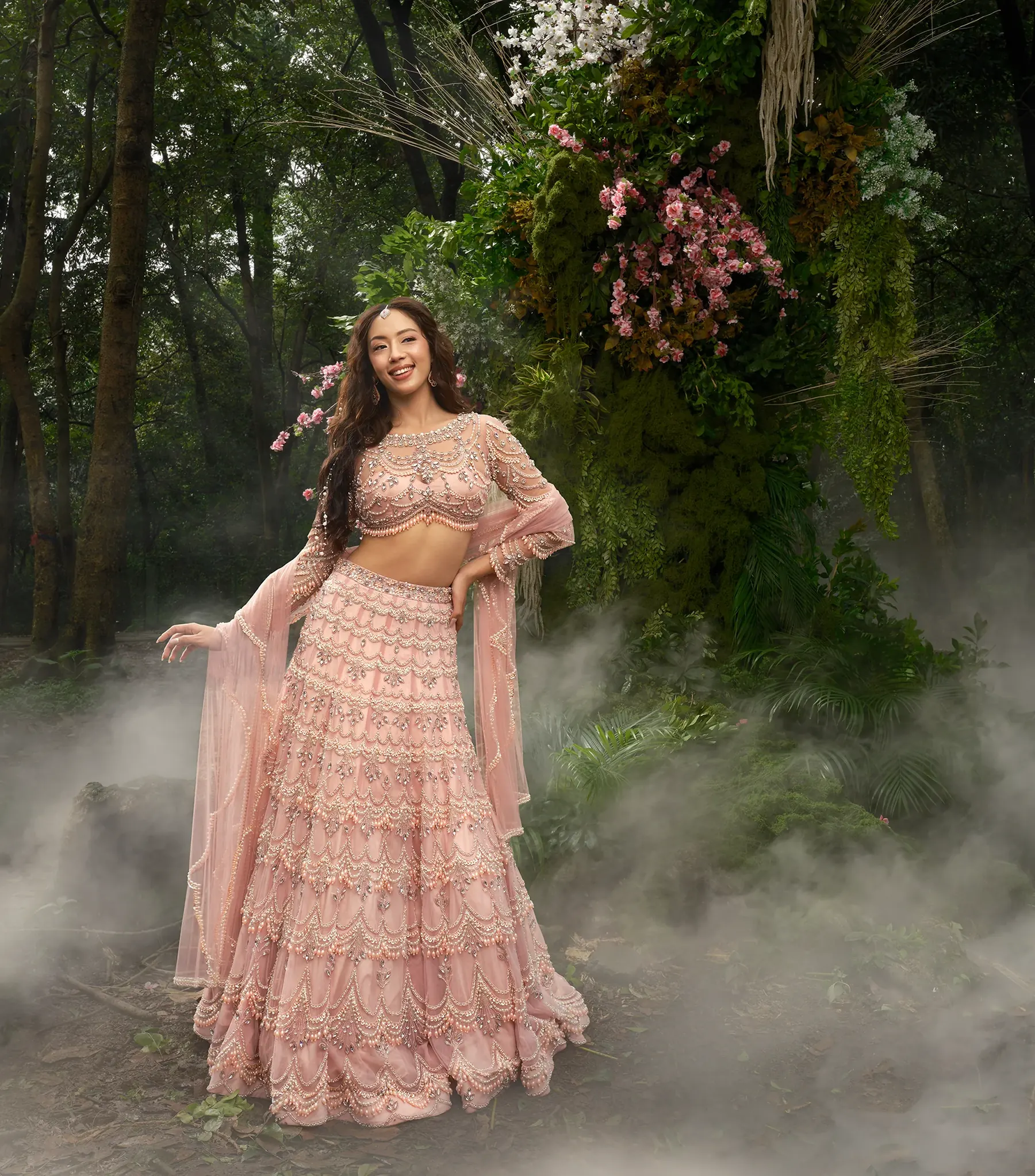A blush pink tiered lehenga with scalloped pearl and bead work and a matching sheer dupatta, beside a floral arch in mist.
