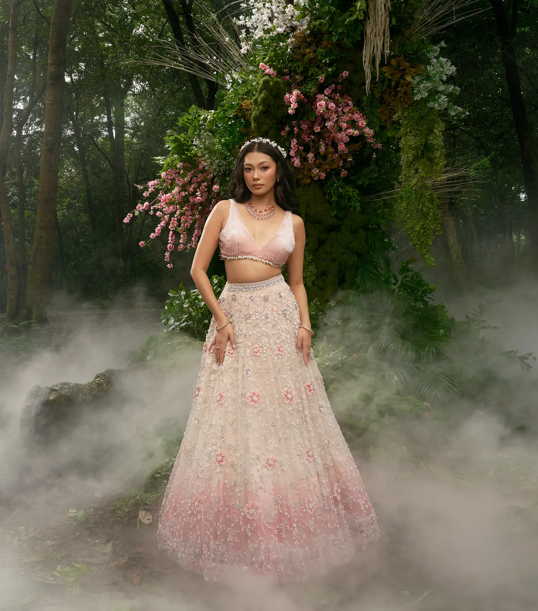 A blush pink lehenga with a beaded bralette blouse and floral embroidery, photographed beneath an arch of pink flowers in mist.