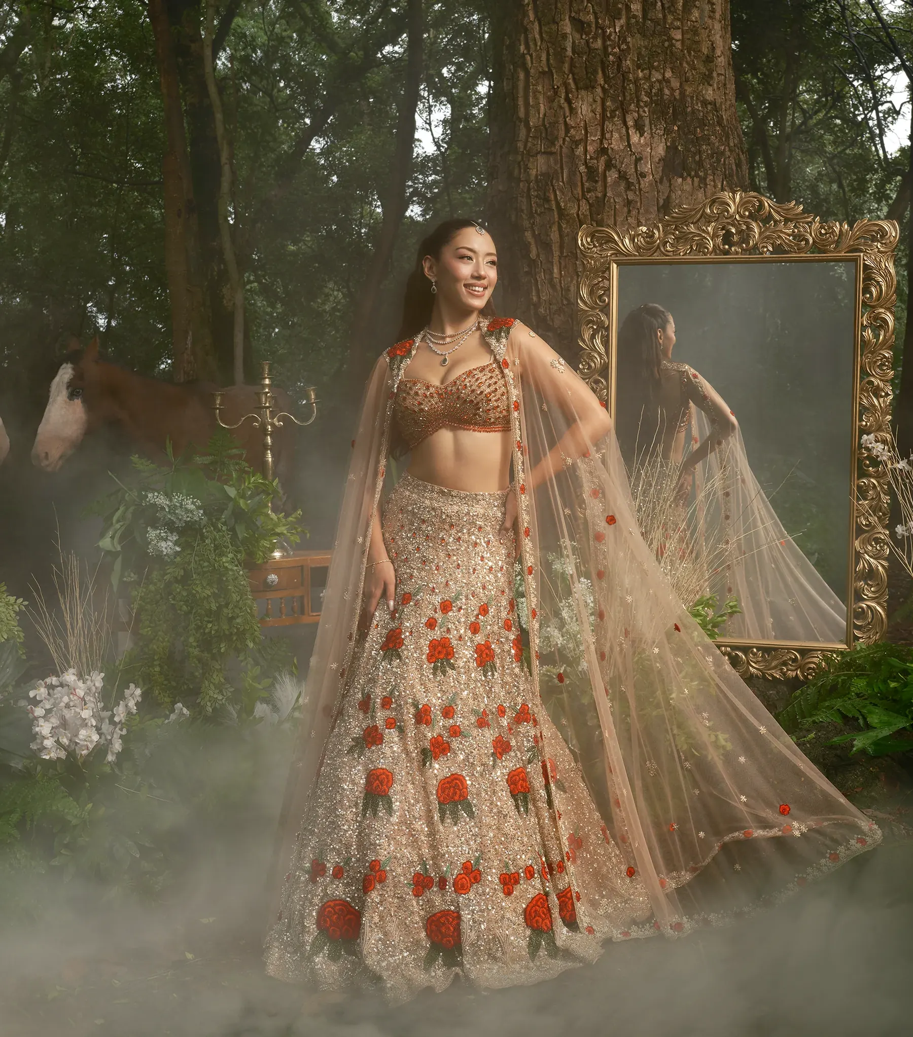 A champagne bridal lehenga with red rose embroidery and a sheer embroidered cape, photographed in mist beside a gilt mirror, lanterns and two horses.