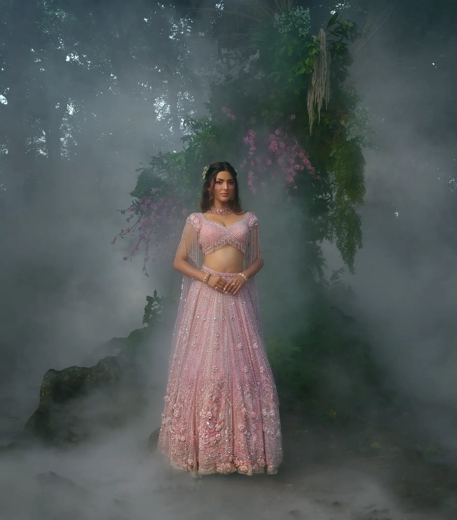 A pale pink lehenga with a beaded, tasselled blouse, standing in thick mist beneath a floral arch.