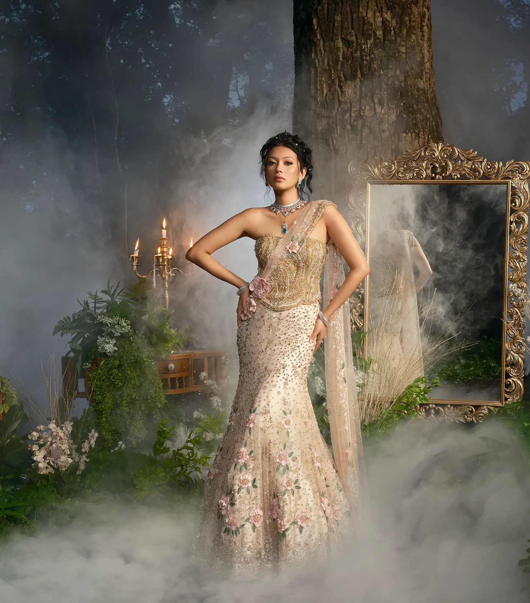 An ivory gown with a gold-embroidered corset, floral appliqué and a sheer drape, photographed beside a gilt mirror and lit candelabra in mist.