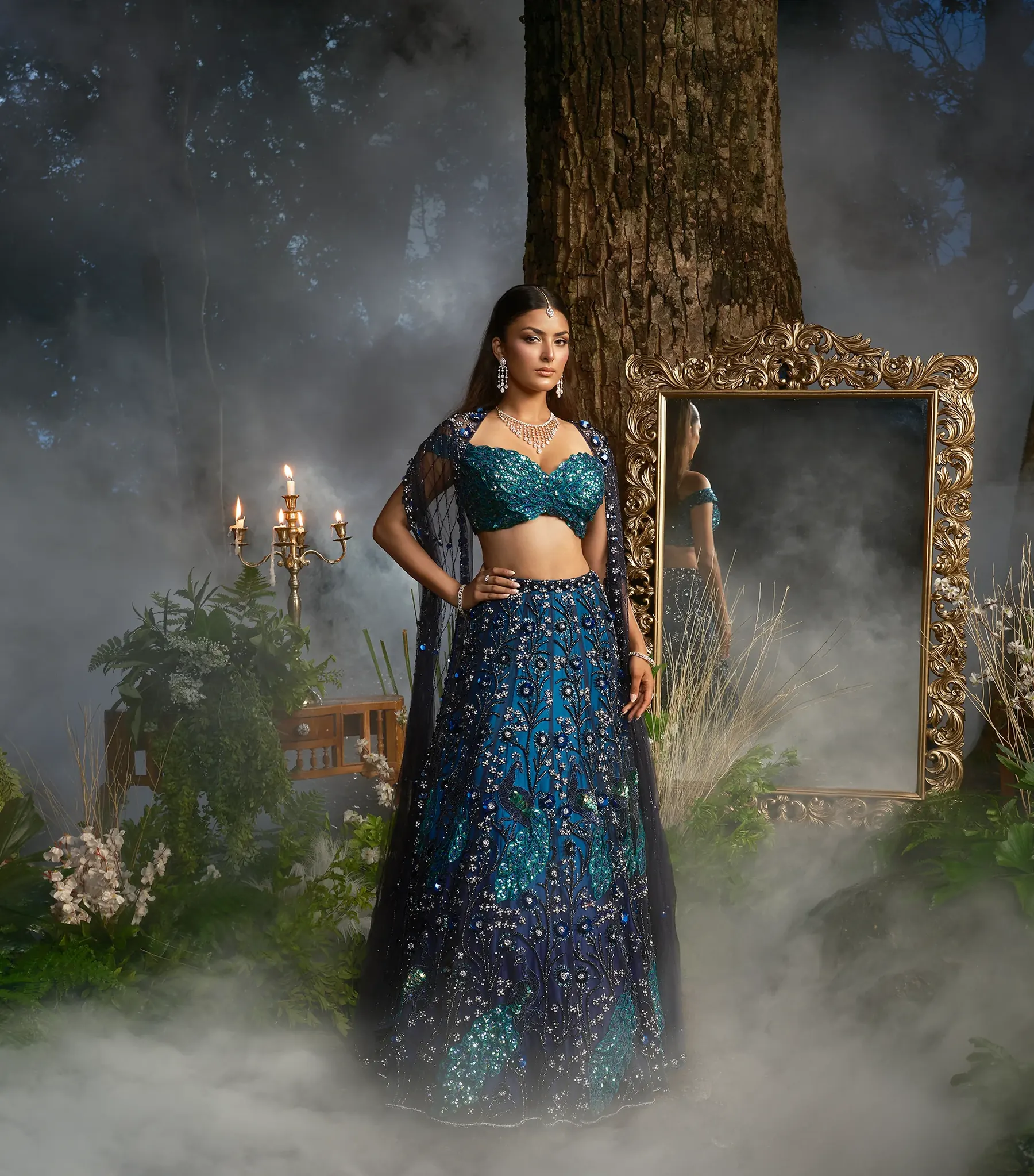 A teal and navy lehenga with sequinned floral embroidery, a beaded blouse and a sheer cape, beside a gilt mirror and lit candles in mist.