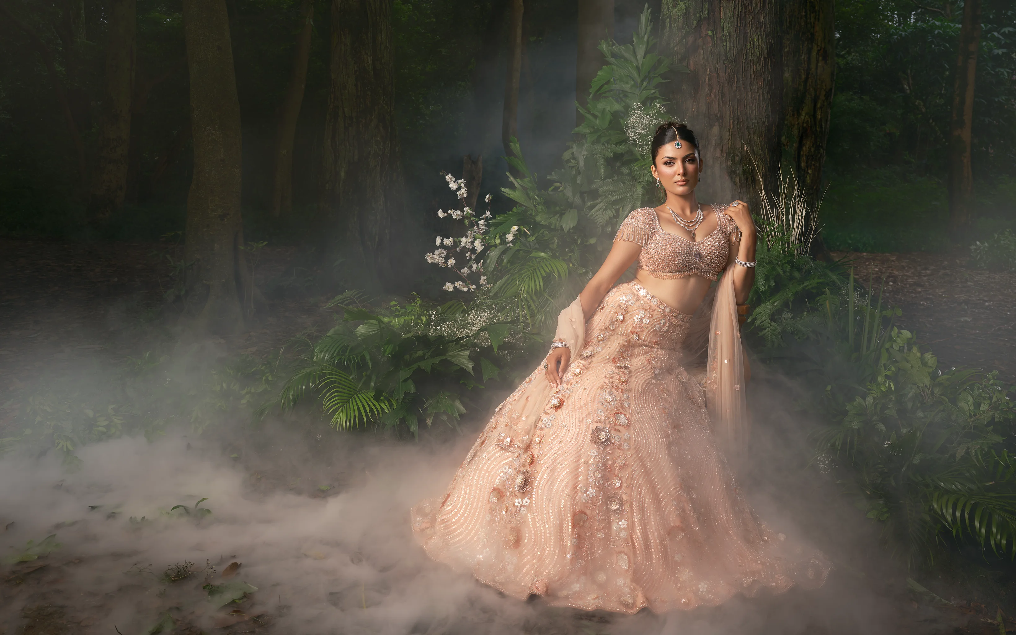 A blush lehenga with floral appliqué and a beaded bralette blouse, photographed beneath a floral arch in mist.