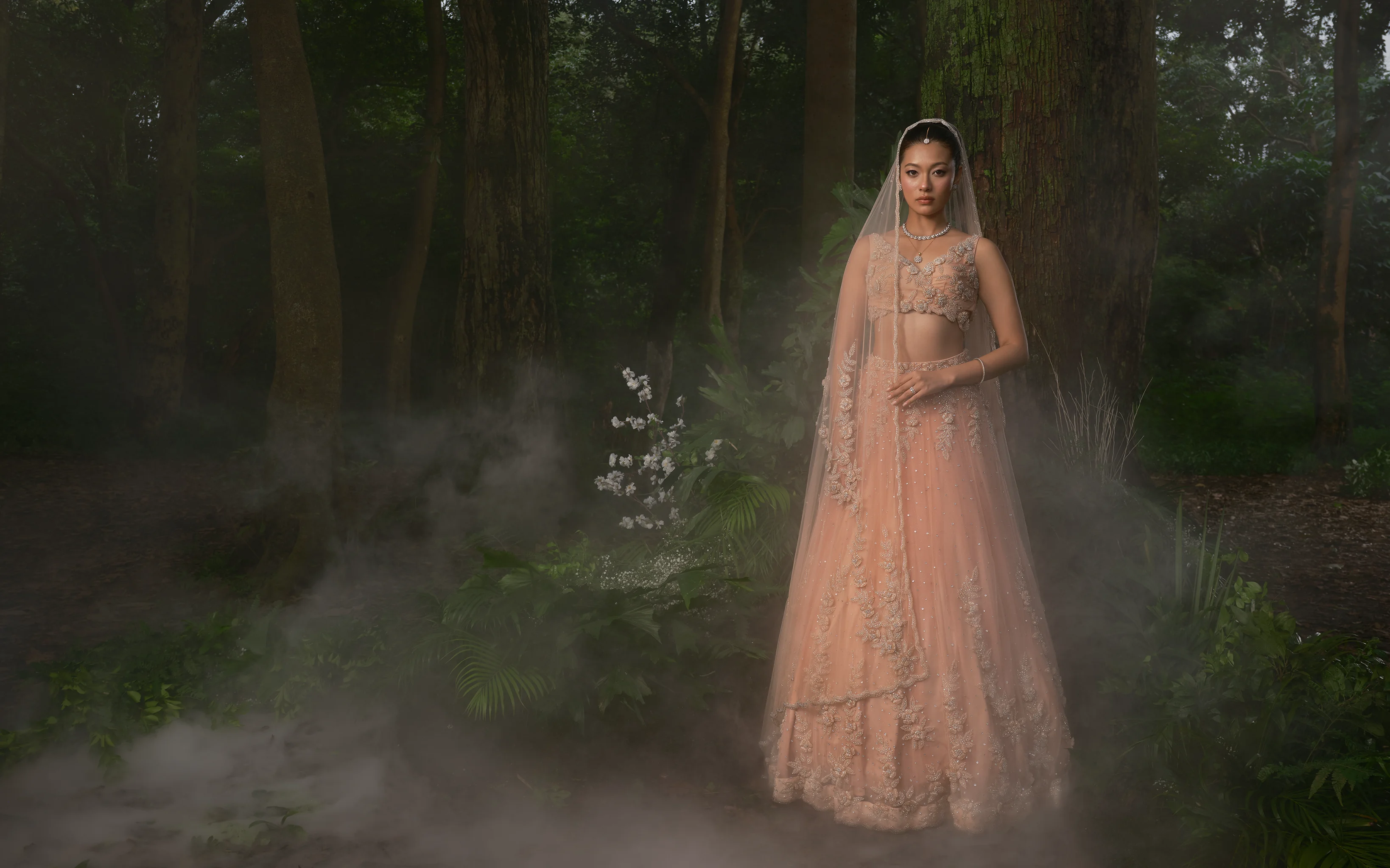 A peach bridal lehenga with a full-length veil and floral appliqué down the drape, in low mist among dark trees.