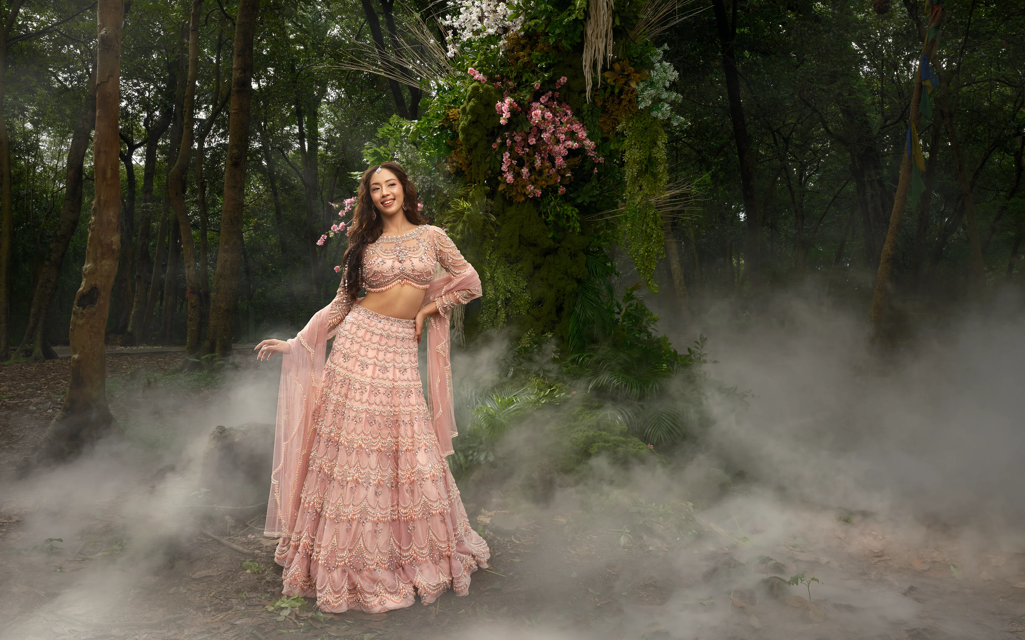 A blush pink tiered lehenga with scalloped pearl and bead work and a matching sheer dupatta, beside a floral arch in mist.